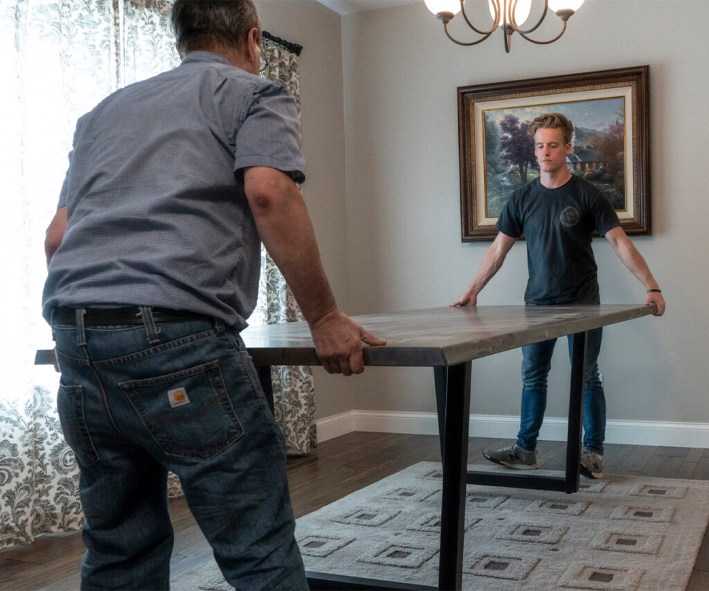 Two people positioning a dining table in a bright dining room.